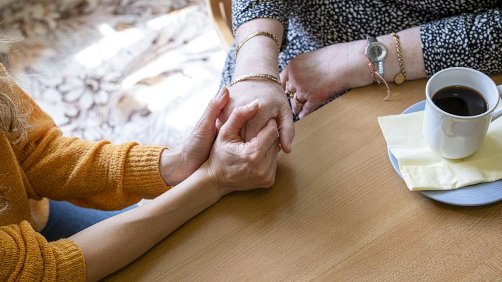 Zu sehen ist ein Bildausschnitt in einem Privatraum. Eine jüngere Frau hält die Hand einer älteren Frau