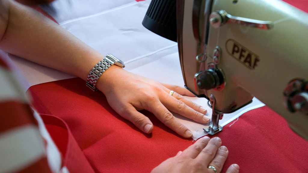 Sur la photo, on voit les mains d'une couturière en train de coudre une croix blanche sur un fond rouge.